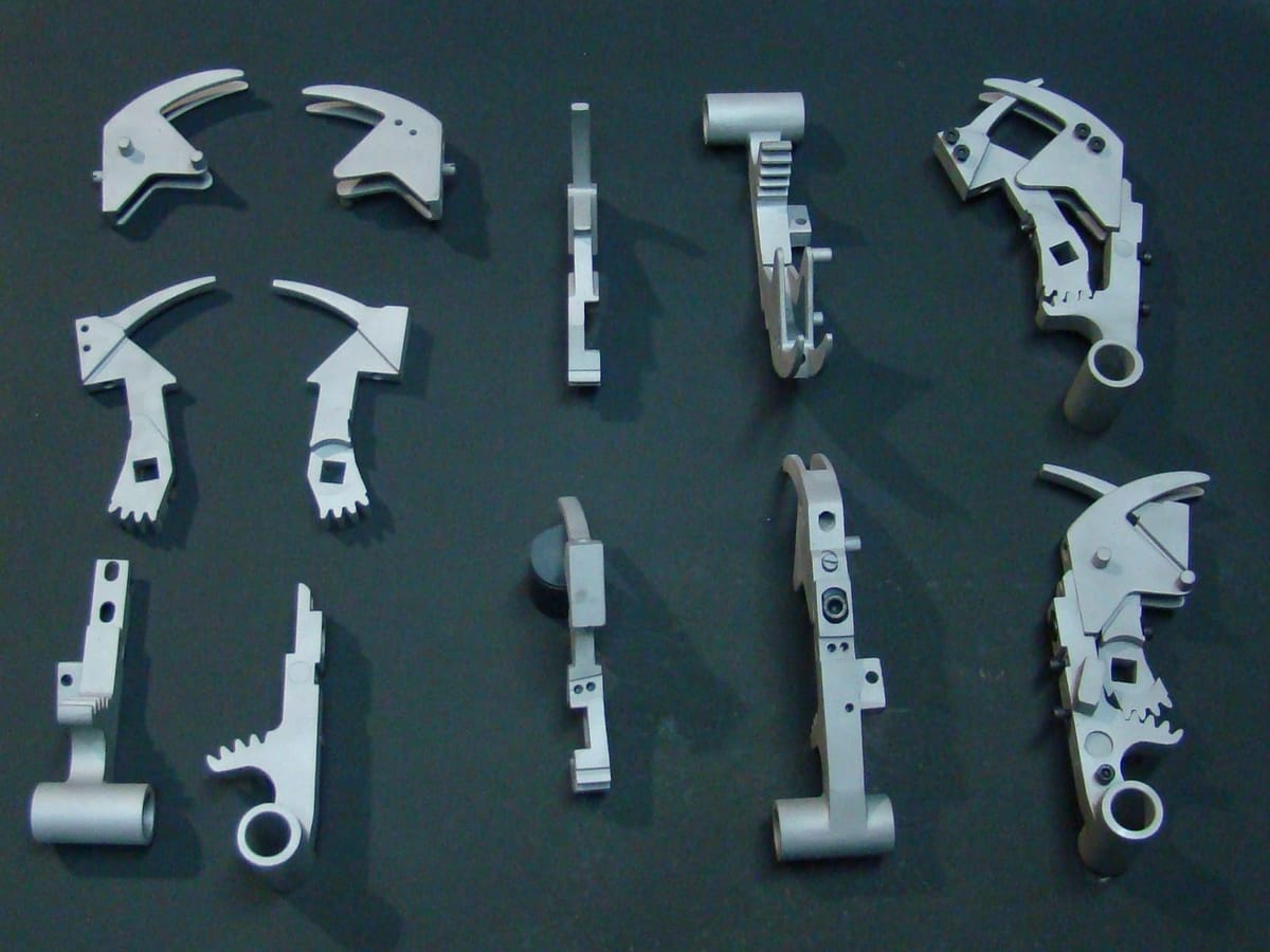 Overhead view of machined aluminum parts on a dark background: arms, cams and toothed rack levers, loose and assembled.